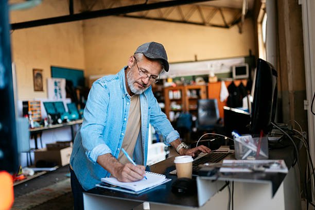 senior small business owner taking notes and working on computer in workshop